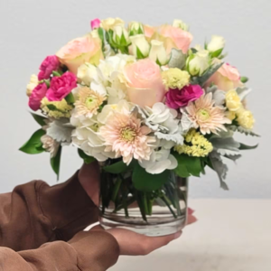 A person holding a bouquet of pink roses and mixed flowers in a glass vase.