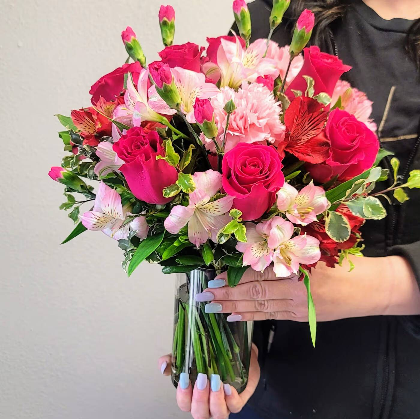 A person holding a bouquet of pink roses, carnations, and alstroemeria in a clear vase.