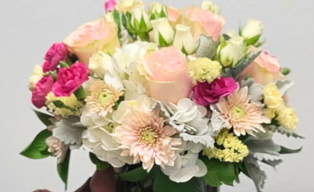 A person holding a bouquet of pink roses and mixed flowers in a glass vase.