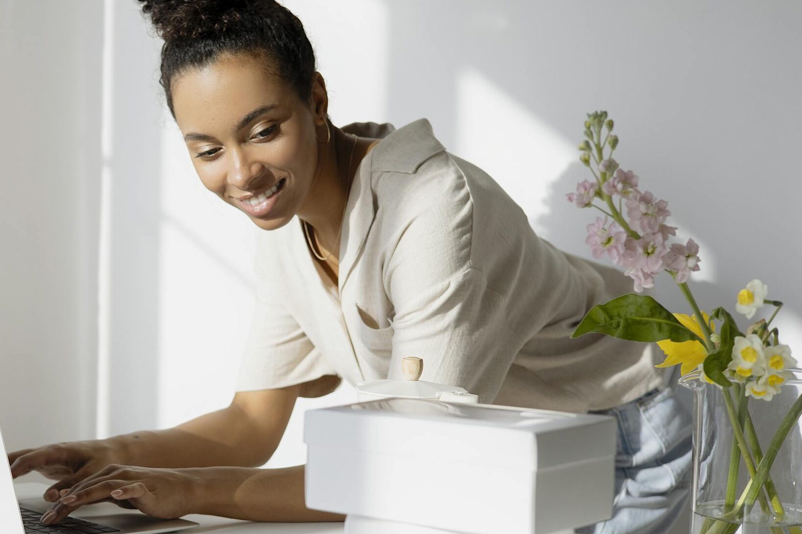 African American woman working on a laptop with boxes and flowers in a bright room.