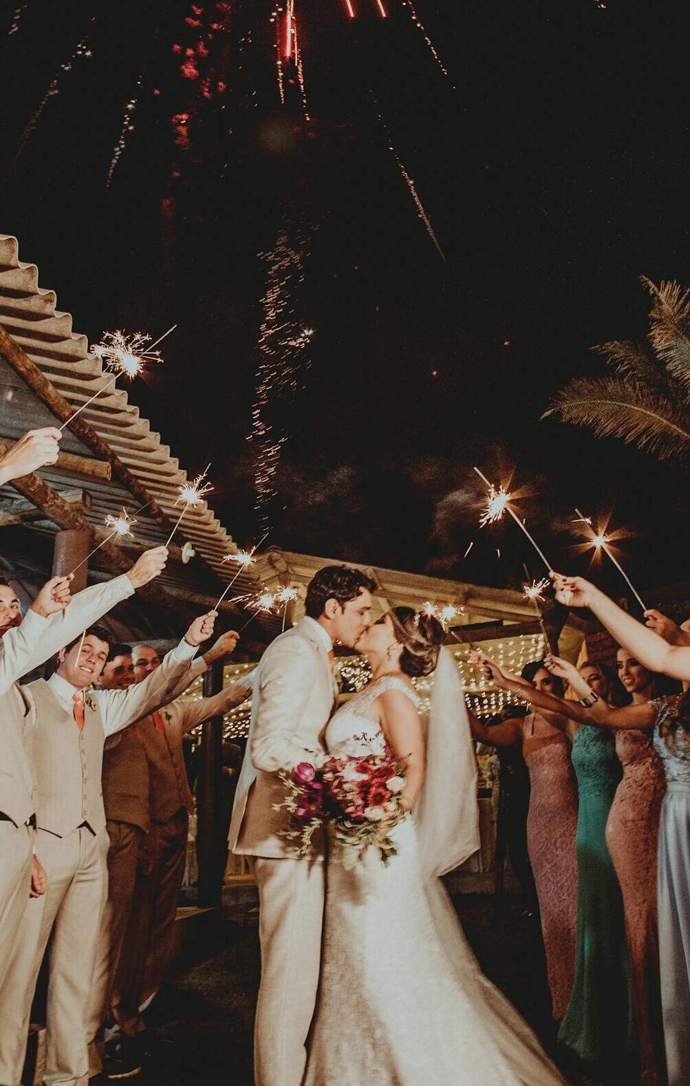 Bride and groom kiss under fireworks surrounded by wedding guests holding sparklers.