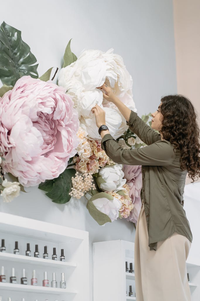 Woman arranging oversized artificial flowers on wall in elegant indoor setting.