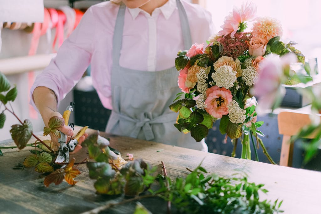 A florist wearing an apron and long sleeves is arranging a vibrant flower bouquet with pruning shears indoors.